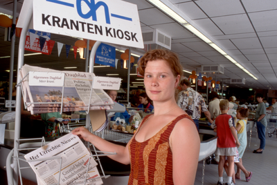 846318 Afbeelding van een jonge vrouw die de krant Het Utrechts Nieuws pakt uit het krantenrek van supermarkt Albert ...